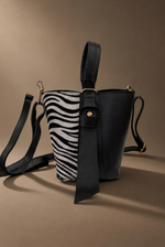 Load image in the gallery viewer,Mala zebra em vista lateral com painel preto e laço dourado, fundo castanho quente
